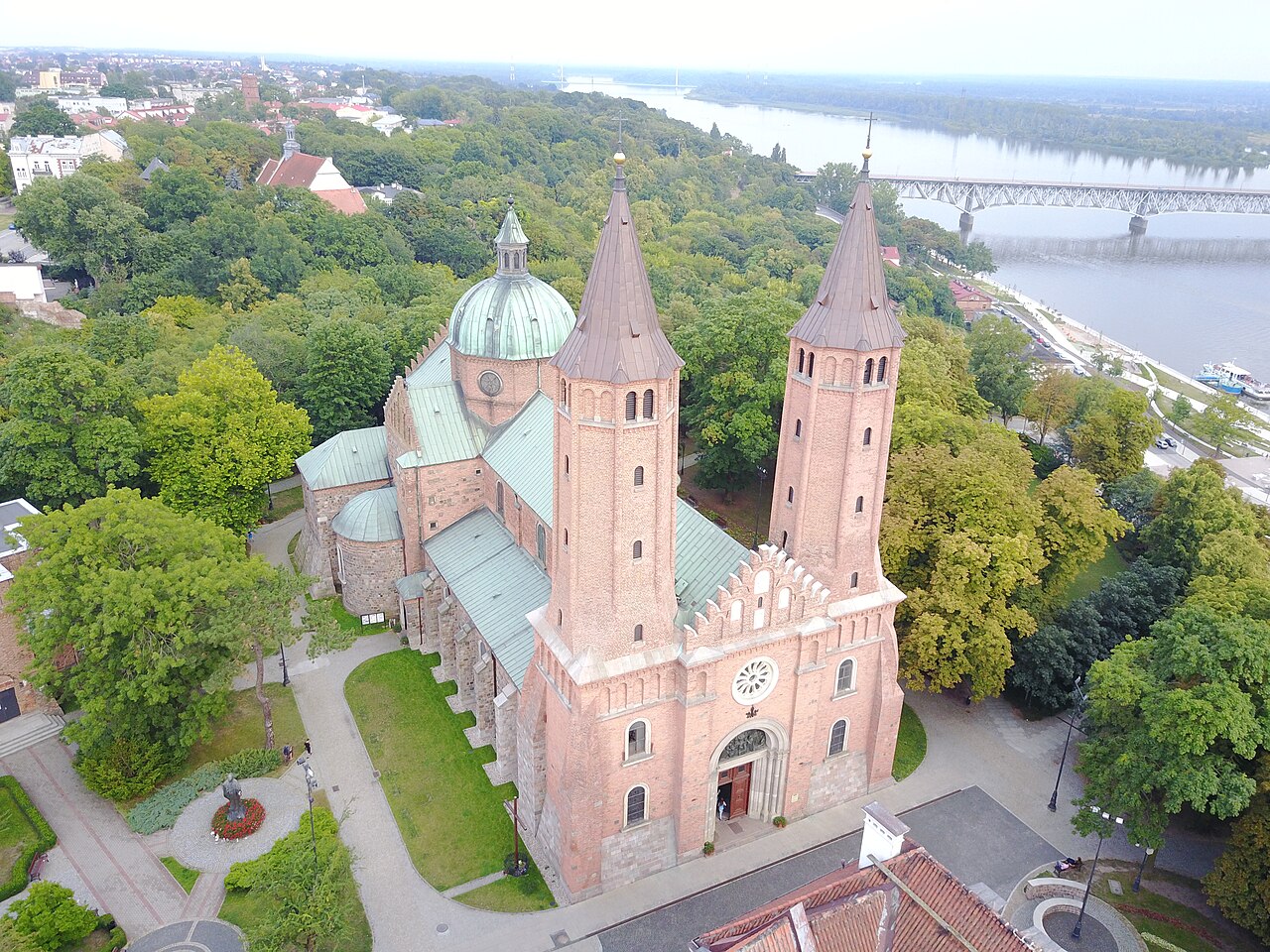 Płock cathedral from the air: a red-brick Romanesque church with two tall towers and a green copper dome, surrounded by trees on a bluff, with the Vistula and a steel bridge behind it