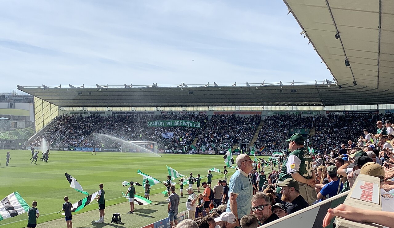 A sunny packed match at Home Park: a large covered grandstand opposite full of supporters with a Green Army banner across it, green and white flags being waved by children along the touchline, pitch sprinklers running and a crowd in green in the foreground