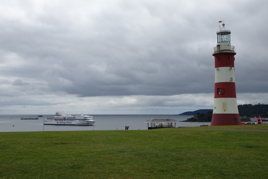 Smeaton's Tower on Plymouth Hoe: a red and white banded stone lighthouse standing on mown grass above the water, with a large white Brittany Ferries ship crossing Plymouth Sound behind it under a grey sky
