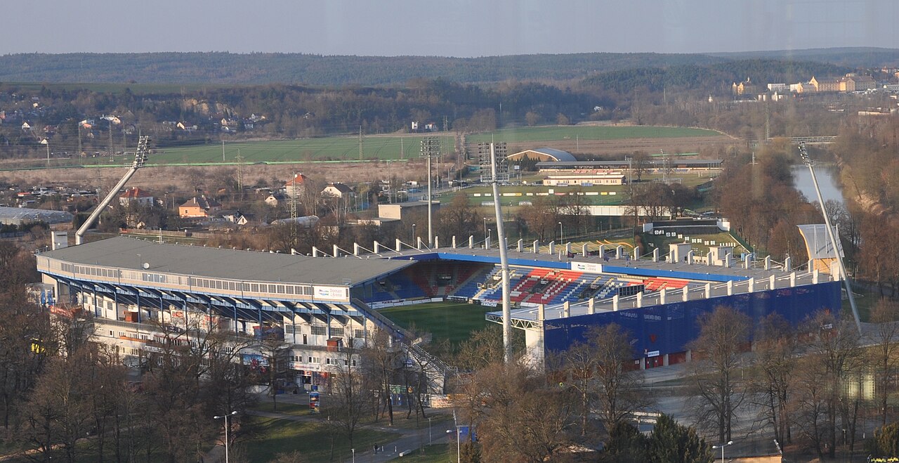 The Doosan Arena in Plzeň seen from a hill above: a compact stadium of mismatched stands in blue and red, standing among bare trees beside a river with fields and woodland behind