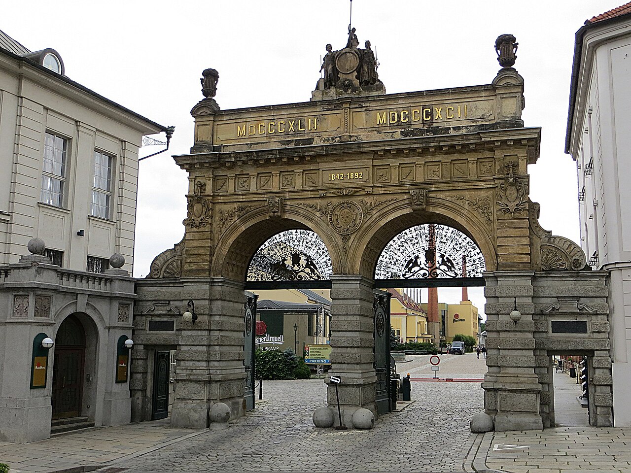 The ornate stone gate of the Pilsner Urquell brewery in Plzeň, a triumphal arch with two arched openings, wrought-iron gates and the dates MDCCCXLII and MDCCCXCII carved across the top