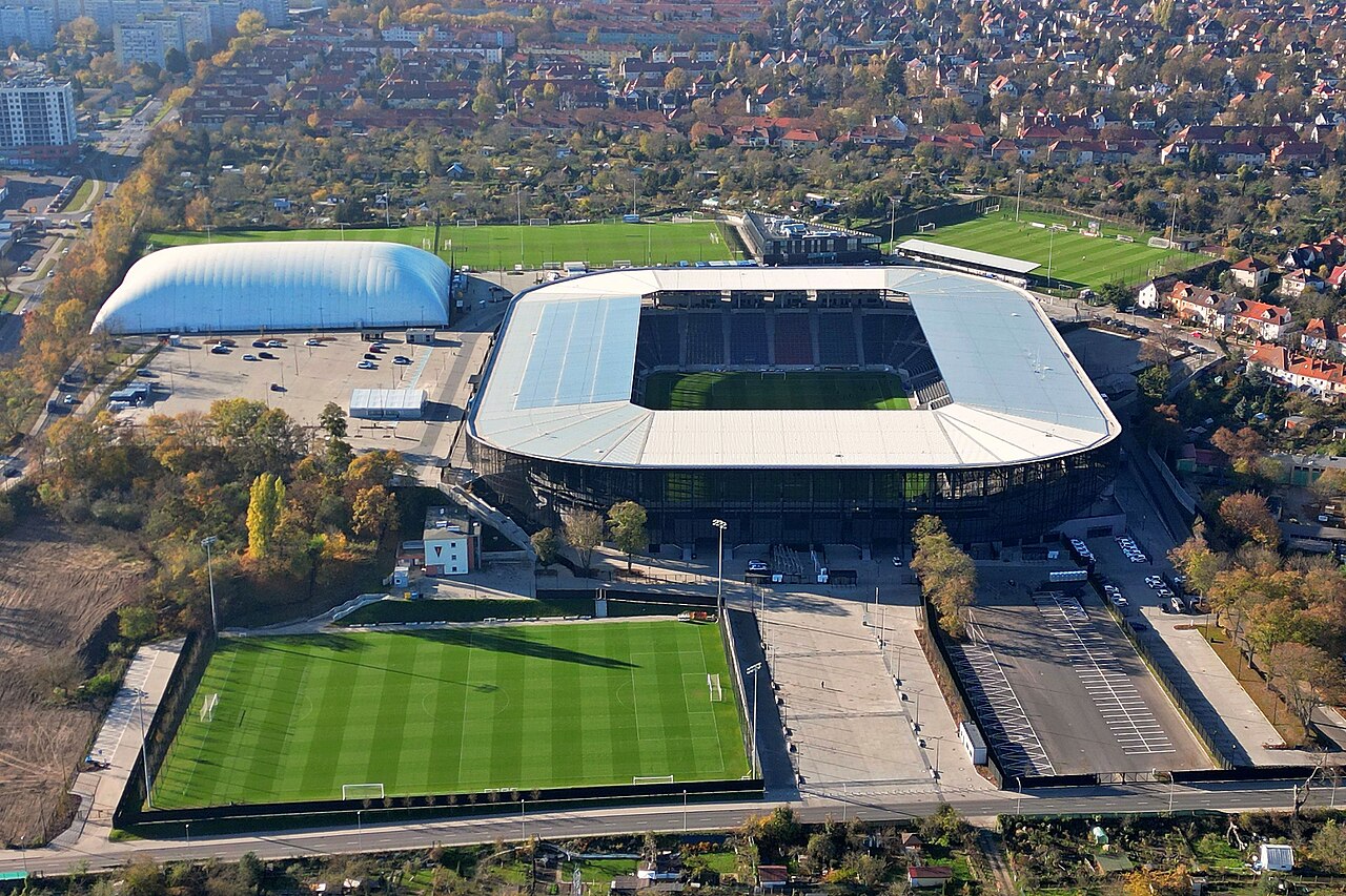 Pogoń Szczecin's rebuilt stadium from the air in autumn: a dark rectangular bowl with a pale roof, training pitches and an inflatable dome beside it, and low suburban housing all around