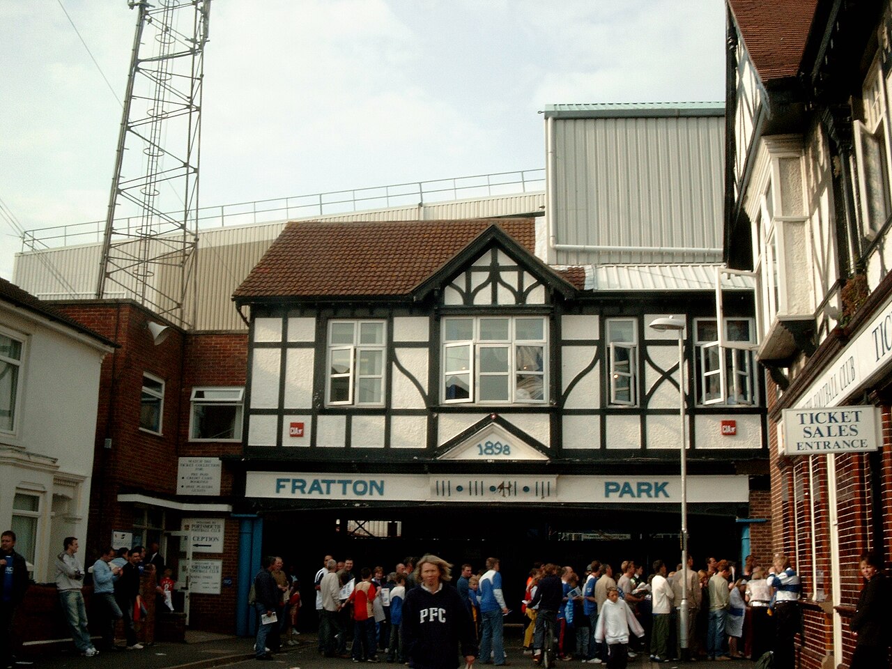 The entrance to Fratton Park: a black and white mock-Tudor gabled building with the date 1898 on it and FRATTON PARK lettered beneath, a crowd of supporters queuing underneath the archway, and a Ticket Sales Entrance sign on the brick building alongside