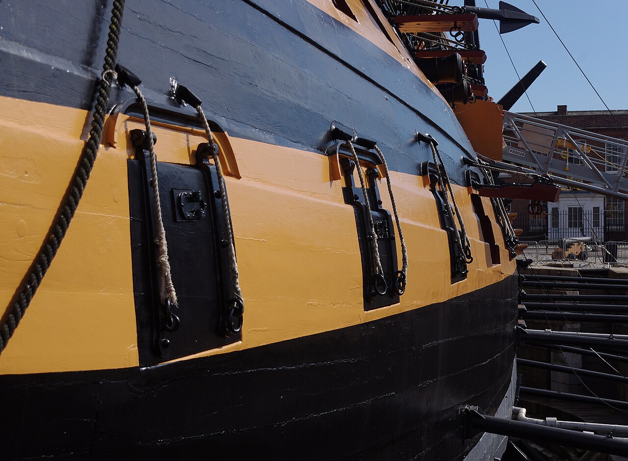The hull of HMS Victory in dry dock, painted in broad black and yellow horizontal bands with square black gun-port lids and heavy iron fittings, ropes running down the side and the ship's rigging visible above