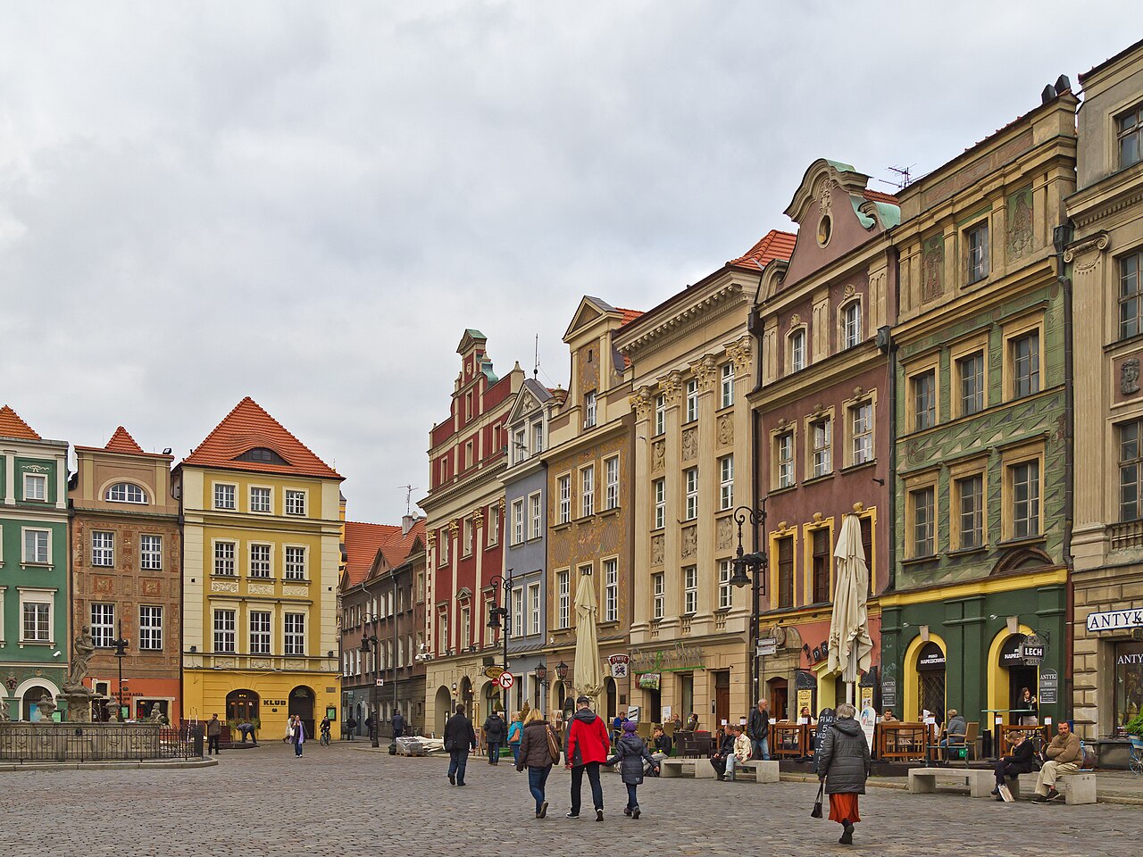 A row of tall, narrow merchant houses in green, yellow, red and ochre along the cobbled Old Market Square in Poznań, with café tables and people walking across the square