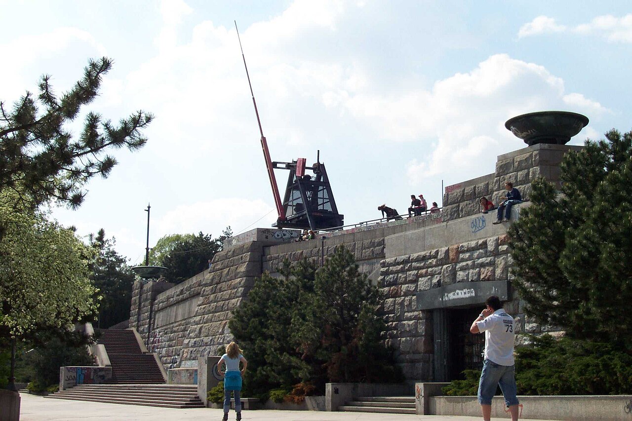 The Prague metronome on Letná: a large red and black metal arm on a massive stone terrace above the city, with people sitting along the edge of the plinth and pine trees to either side