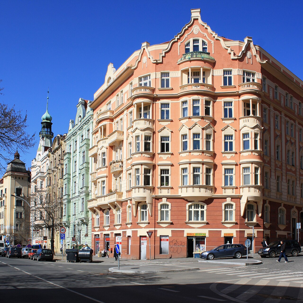 A salmon-pink Art Nouveau corner tenement in Vršovice, Prague, with bay windows and a decorated gable, ordinary shops at street level and a green church tower visible along the street behind