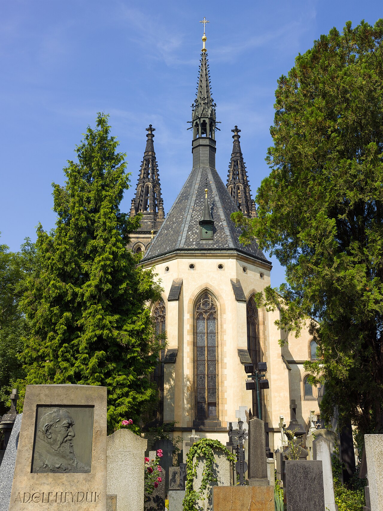The Gothic basilica of St Peter and St Paul at Vyšehrad in Prague, its twin spires rising behind cypress trees, seen from the cemetery with carved headstones and a bronze portrait plaque in the foreground
