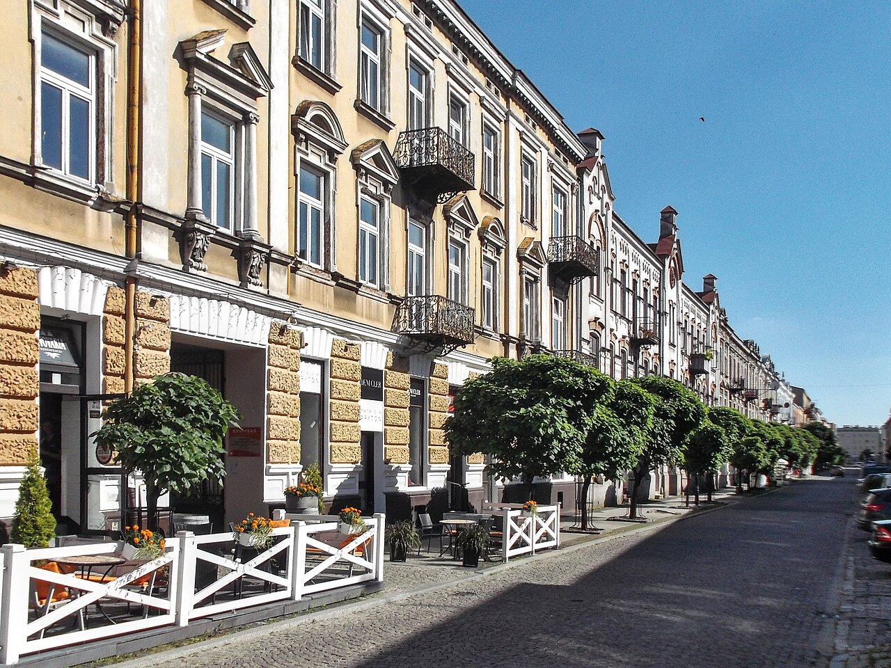 A street in central Radom: a terrace of ornate nineteenth-century townhouses in yellow and white with wrought-iron balconies, trimmed trees and café tables along the cobbled pavement