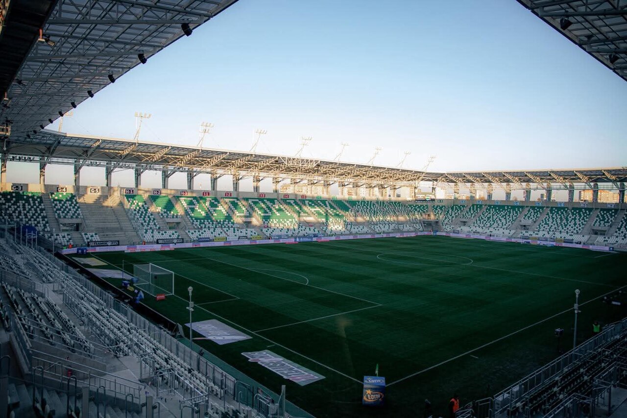 The inside of Radomiak's rebuilt stadium at sunset, seen from a corner: empty green and white seats under a covered roof, sector letters on the stand, a striped pitch and low evening light across it