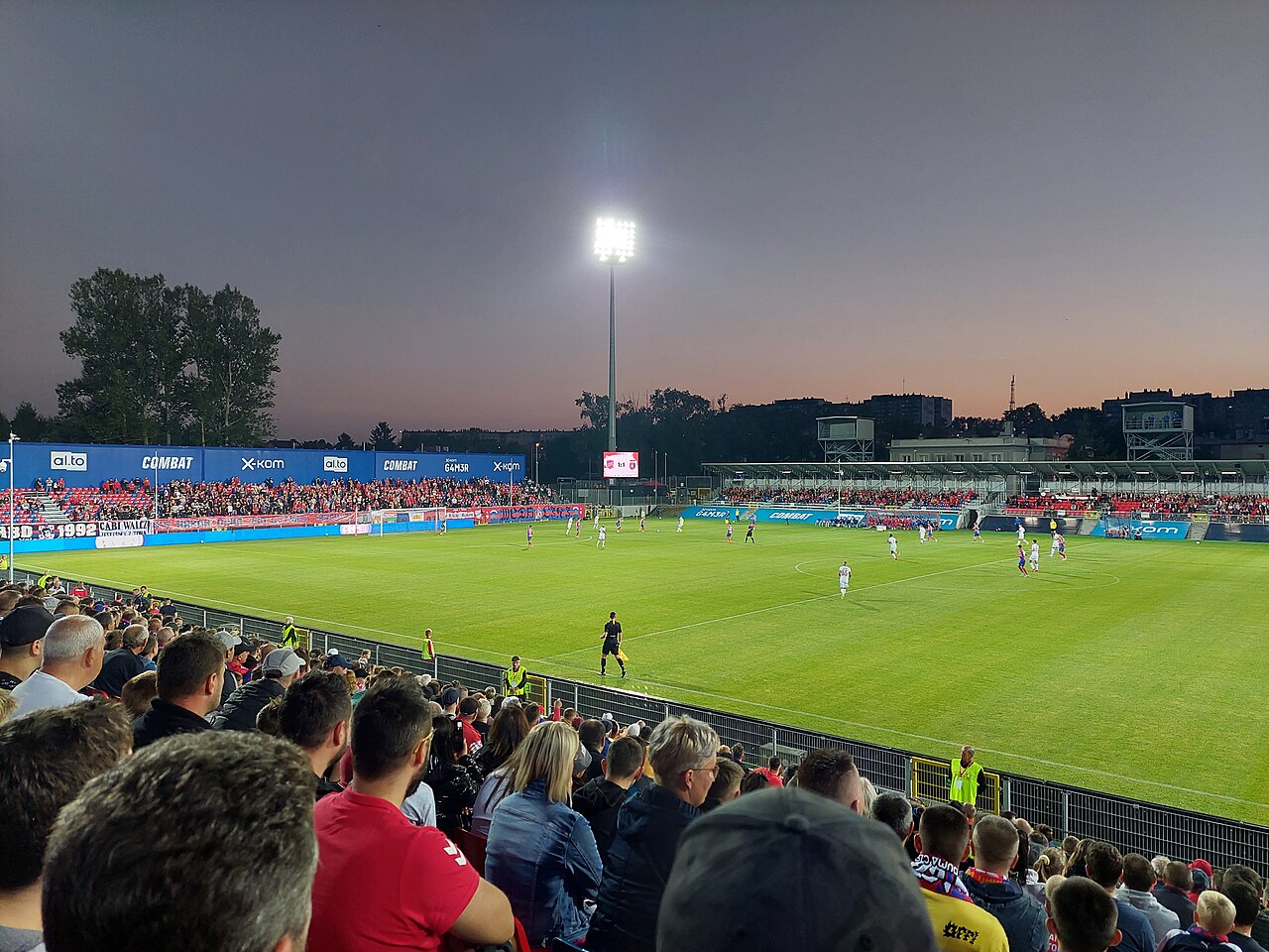 A night match at Raków's small stadium seen from the stand: floodlight pylon against a dusk sky, packed low stands in red and blue on both sides, houses visible over the far touchline