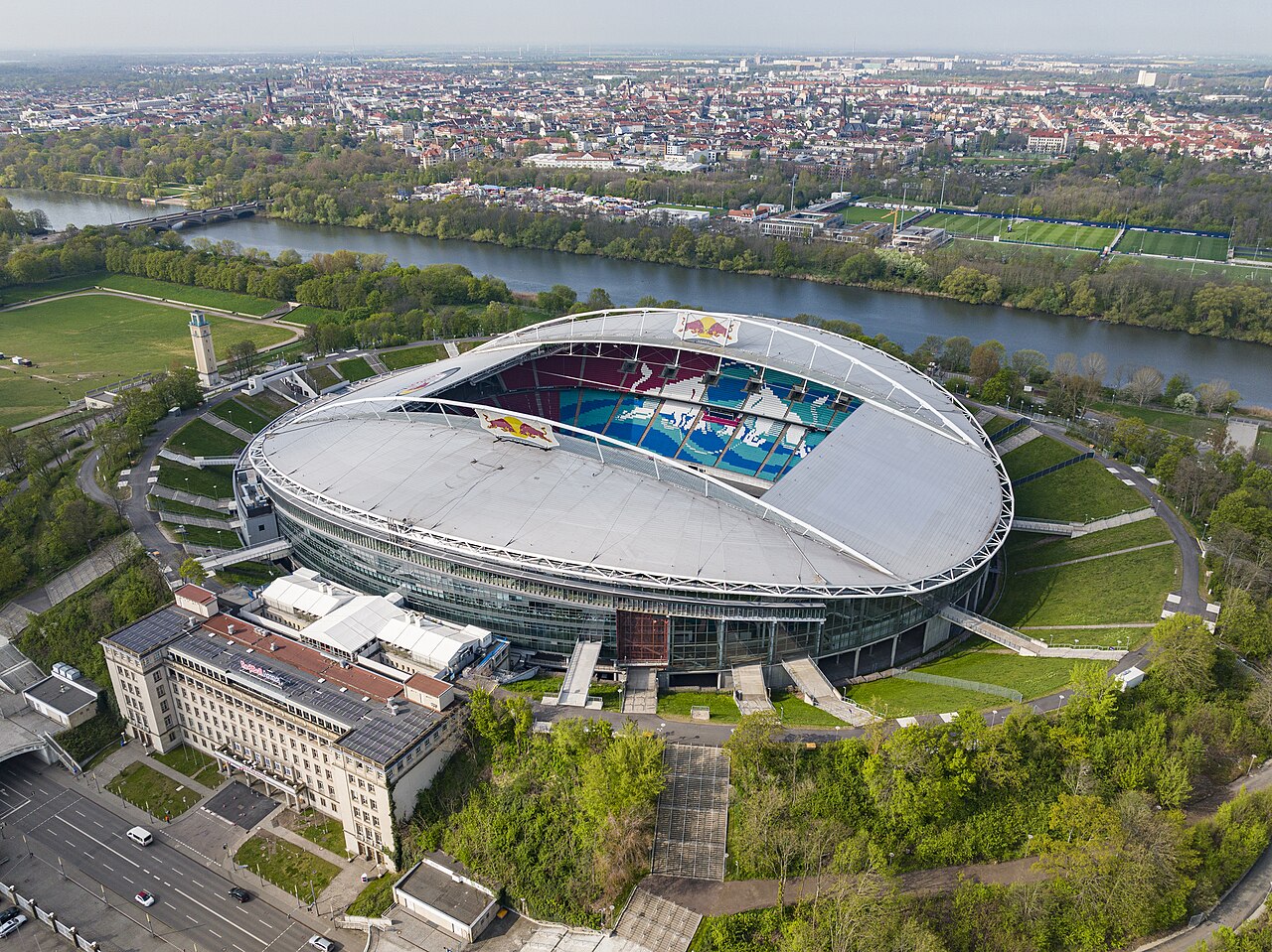 The Red Bull Arena from the air: an oval stadium with a white roof sitting inside the grass embankments of a much larger older bowl, with a river and the Leipzig skyline behind