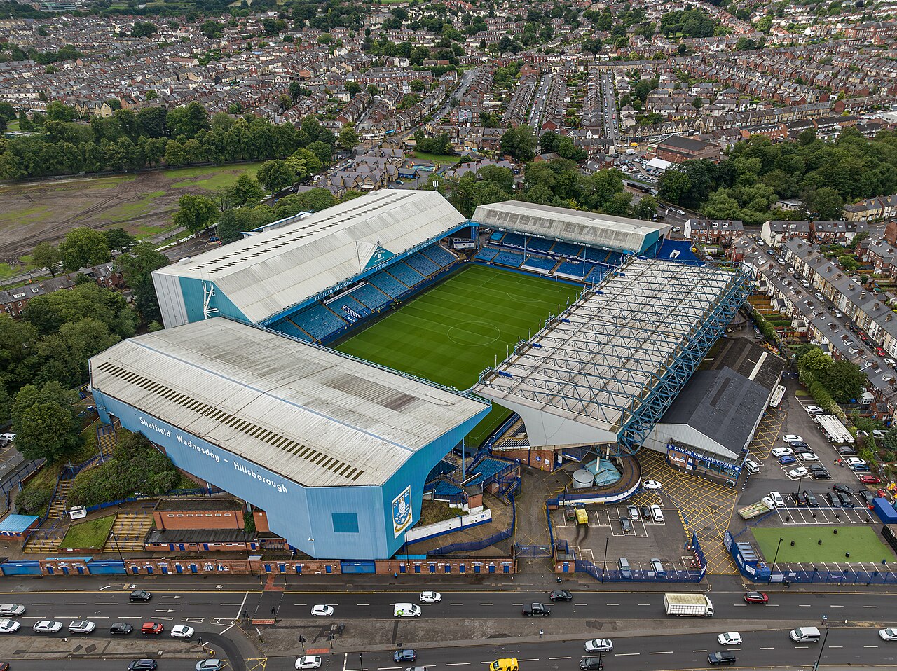 Hillsborough from the air: a blue and white four-stand football ground with Sheffield Wednesday Hillsborough painted along one roof, a large blue steel cantilever stand on one side, a main road along the front and streets of terraced housing pressing right up against the other three sides