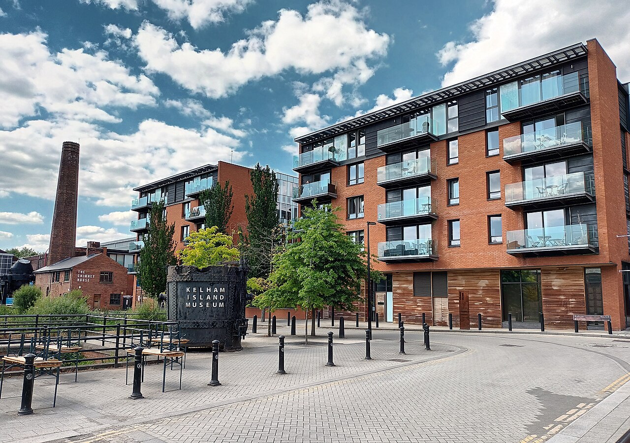The Kelham Island quarter in Sheffield: a large black cast-iron sign reading KELHAM ISLAND MUSEUM on a paved square, with a tall brick industrial chimney and an old brick building on one side and new red-brick apartment blocks with glass balconies on the other
