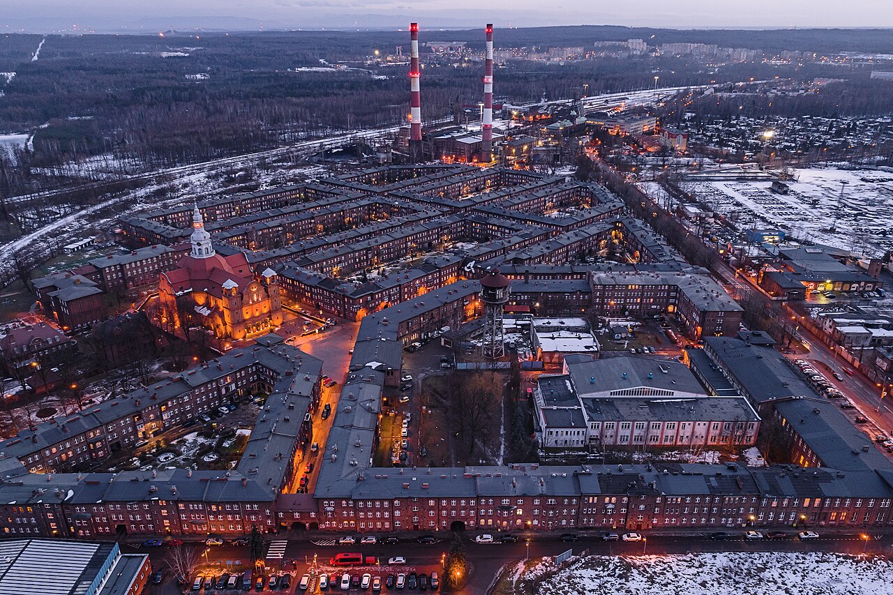 Aerial view at dusk of Nikiszowiec in Katowice: dense blocks of red-brick miners' housing arranged around courtyards, a church lit orange, and two striped colliery chimneys on the horizon