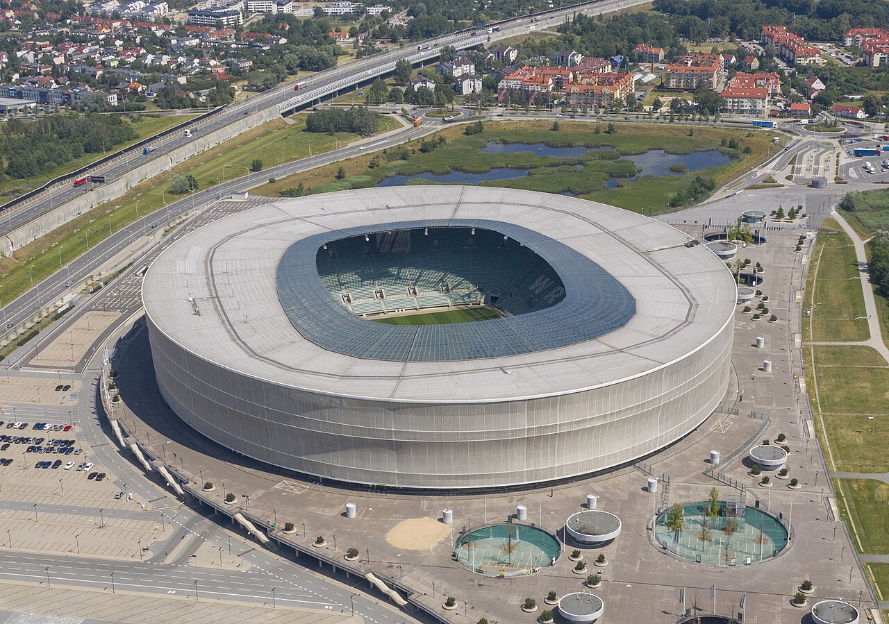 The Tarczyński Arena from the air: a large oval stadium wrapped in a perforated white shell, standing beside a motorway with suburban housing and ponds around it