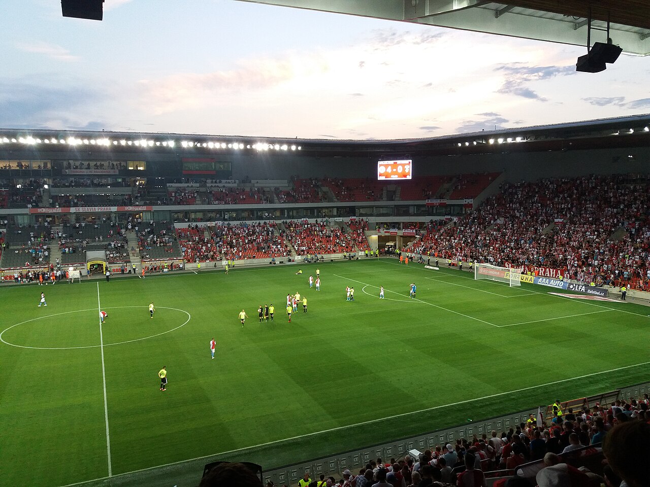 A match at Slavia Prague's stadium at dusk seen from a corner: red seats filled with supporters in four single-tier covered stands, a floodlit green pitch and SK SLAVIA PRAHA on the advertising boards
