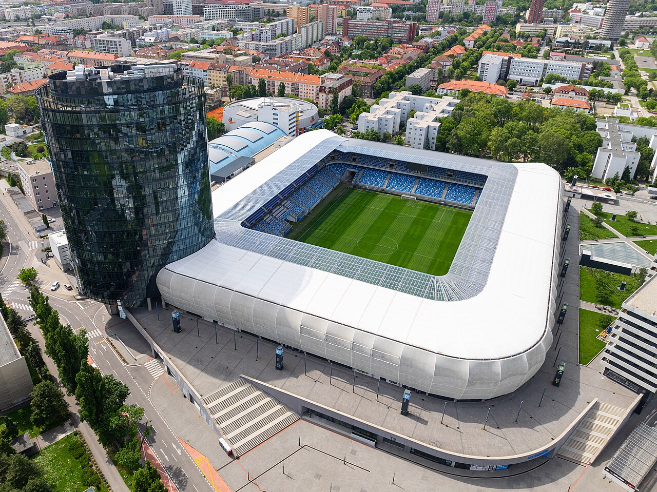 Tehelné pole from the air: a rectangular stadium wrapped in white cladding with blue seats inside, a cylindrical glass office tower beside it and Bratislava's apartment blocks all around