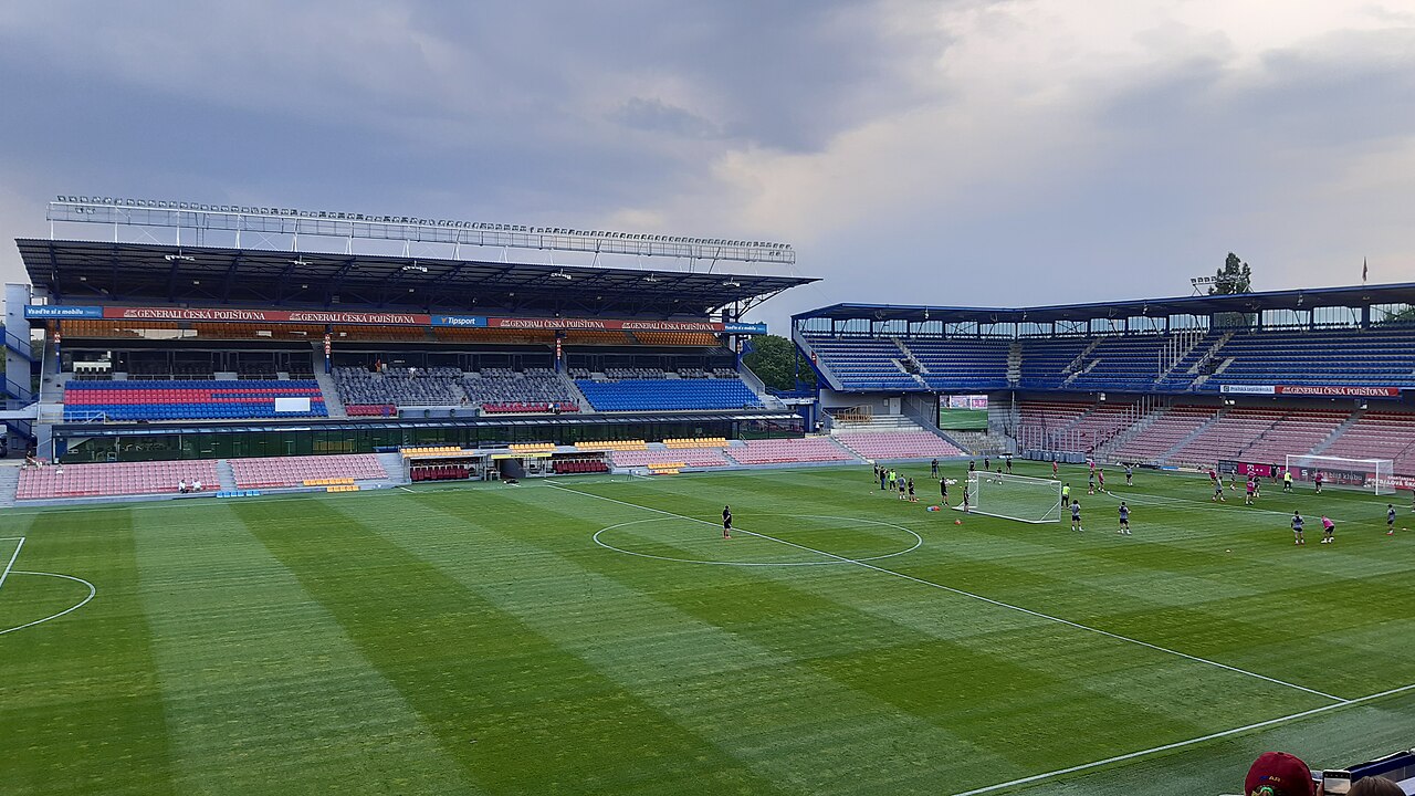 Letná stadium empty before a match: two stands meeting at a corner, seats in red, blue and white, a covered main stand on the left and players warming up on the striped pitch under a grey sky