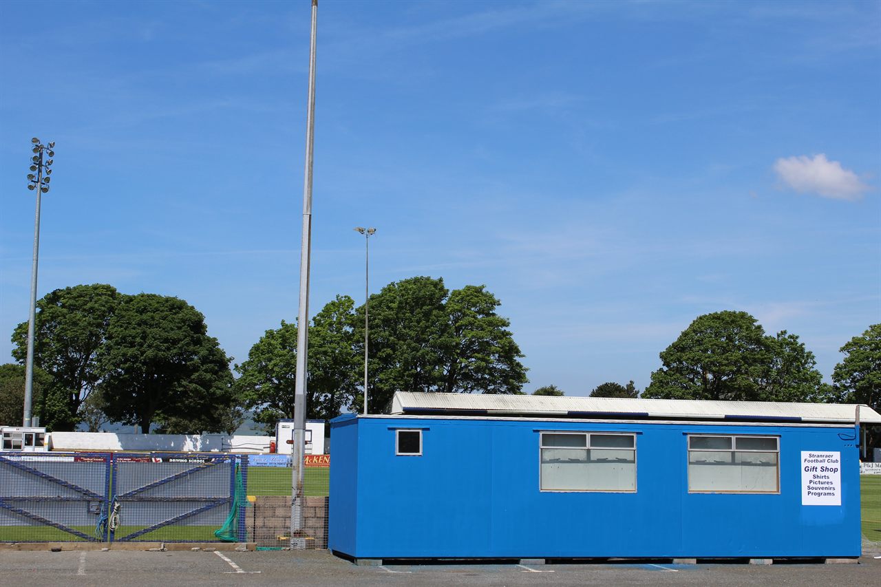 A small single-storey club gift shop at a lower-league ground, painted in club colours with a modest sign, next to the perimeter fence of the stadium