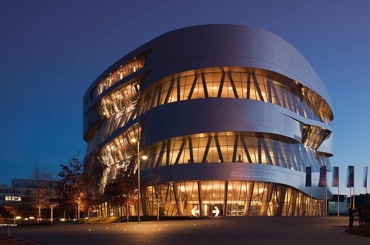 The Mercedes-Benz Museum at dusk: a silver building of curved overlapping bands with glass slots between them, lit warmly from inside, standing on an empty paved forecourt