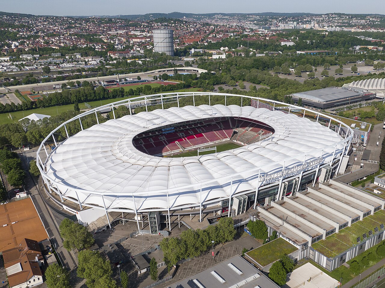 The Stuttgart stadium from the air: an oval bowl of red seats under a white tented roof ring, with a sponsor name across the facade and the city rising up wooded slopes behind