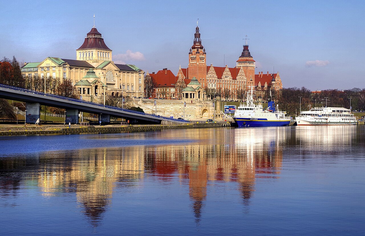 The Chrobry Embankment in Szczecin seen across the Oder at sunset: a monumental terrace with a domed museum and a red-brick tower, ships moored below, everything reflected in the water