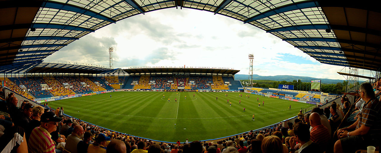 A wide view inside Na Stínadlech during a match: four covered stands filled with supporters in yellow and blue, a floodlit green pitch, and the wooded ridge of the Ore Mountains on the horizon beyond the far stand