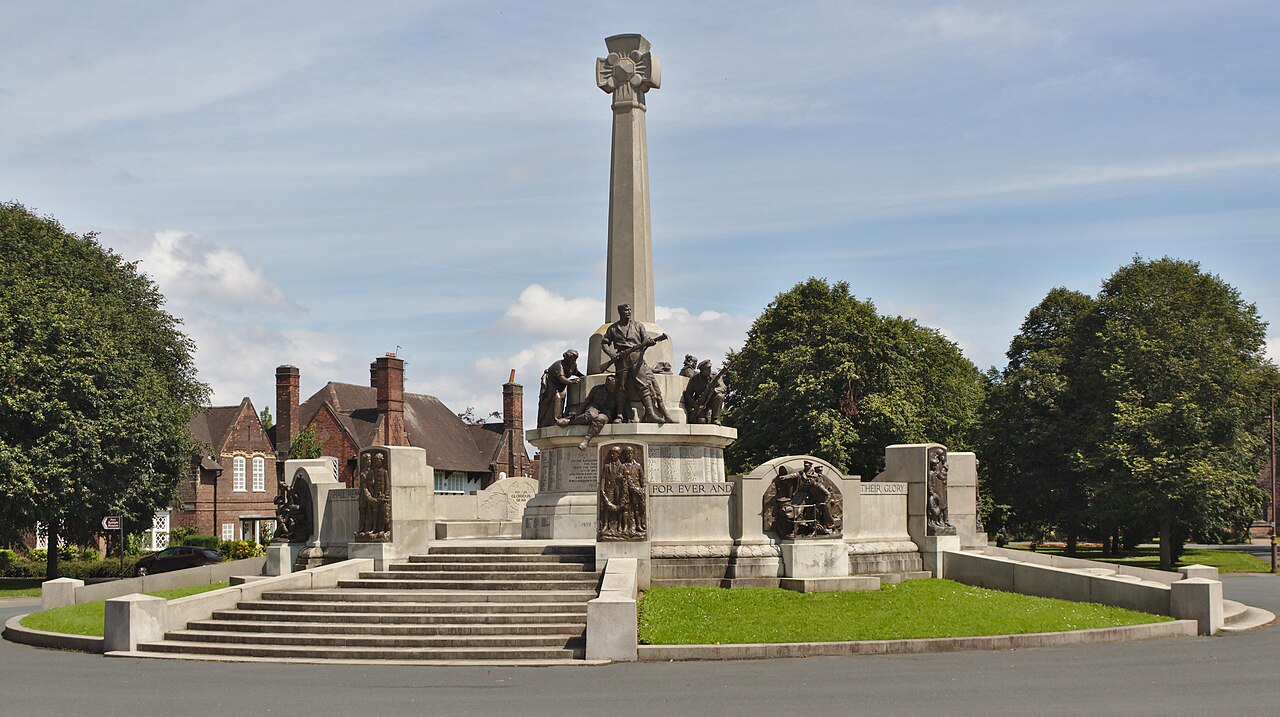 The war memorial at Port Sunlight: a tall stone cross on a column above a granite plinth crowded with bronze figures of soldiers and civilians, standing on a wide flight of steps in the middle of a green, with half-timbered village houses and mature trees behind