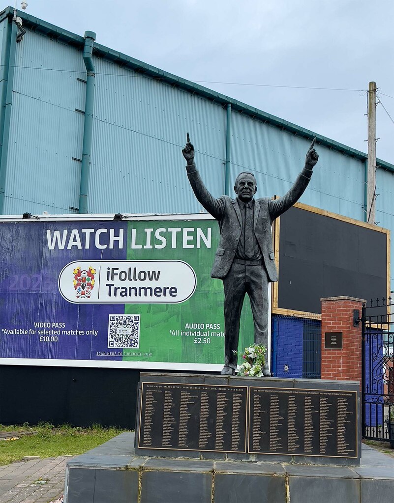 The Johnny King statue outside Prenton Park: a bronze figure of a man in a suit with both arms raised and index fingers pointing skywards, standing on a plinth covered with hundreds of engraved names, with the green corrugated wall of a stand and a club hoarding behind