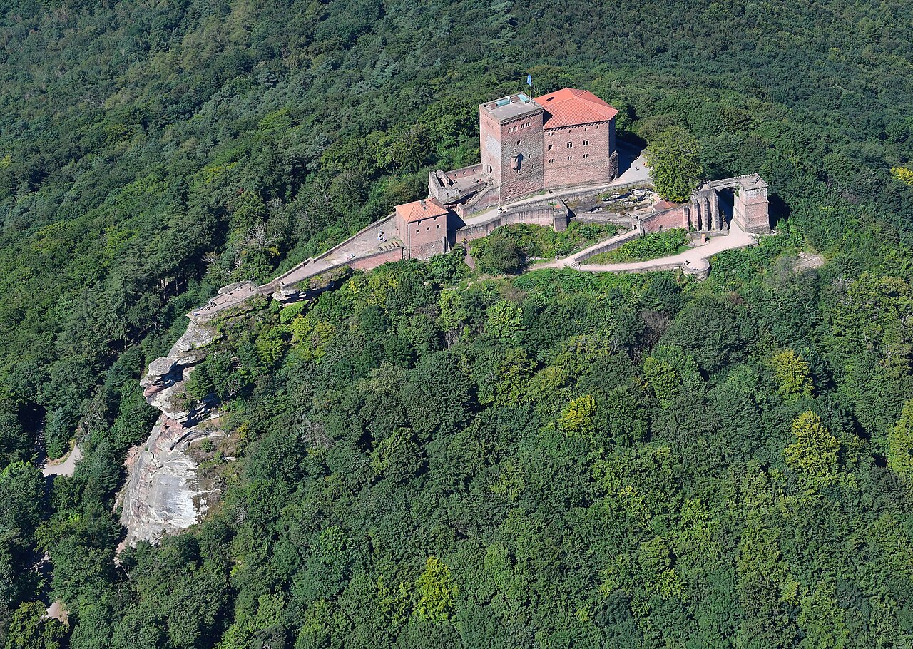 Trifels Castle from the air: a red sandstone castle with a tall keep and red tiled roofs standing on a narrow rock spur, ruined outbuildings along the ridge, and dense forest covering the hills in every direction