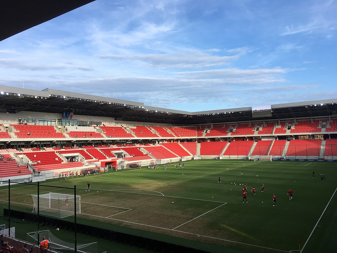 Inside the Anton Malatinský Stadium: banks of red seats in two tiers under a wide flat roof, players warming up on the pitch, and the corner where two stands meet