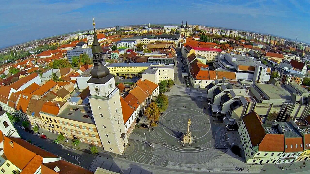 Trnava from the air: the town hall tower and a gilded plague column on the paved main square, red-roofed old town all around, and the angular grey roofs of the stadium and arena complex on the right