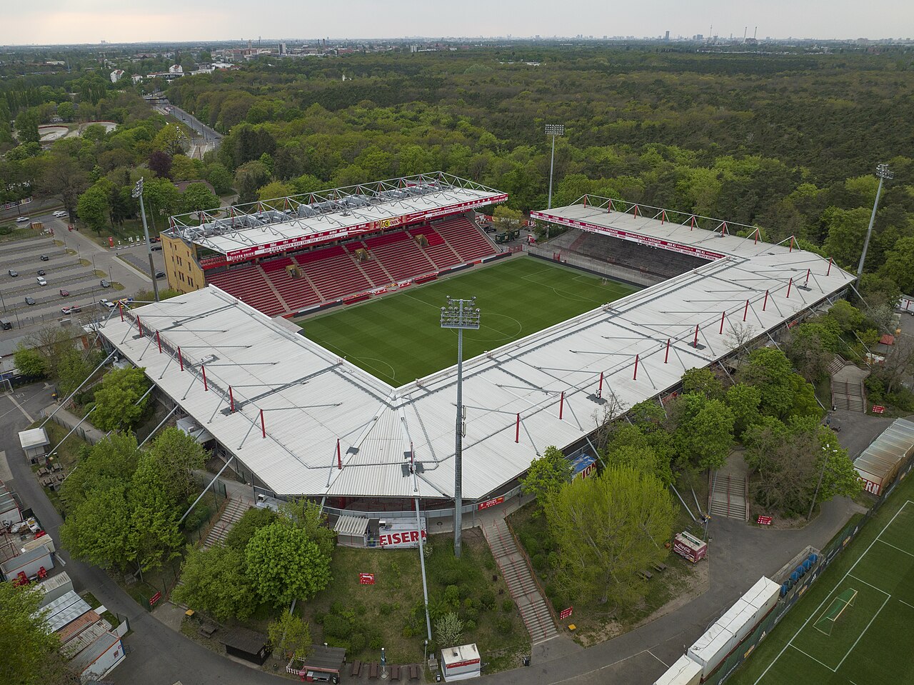 An der Alten Försterei from the air: a small stadium surrounded on all sides by forest, with one red seated stand and three low covered terraces around a green pitch