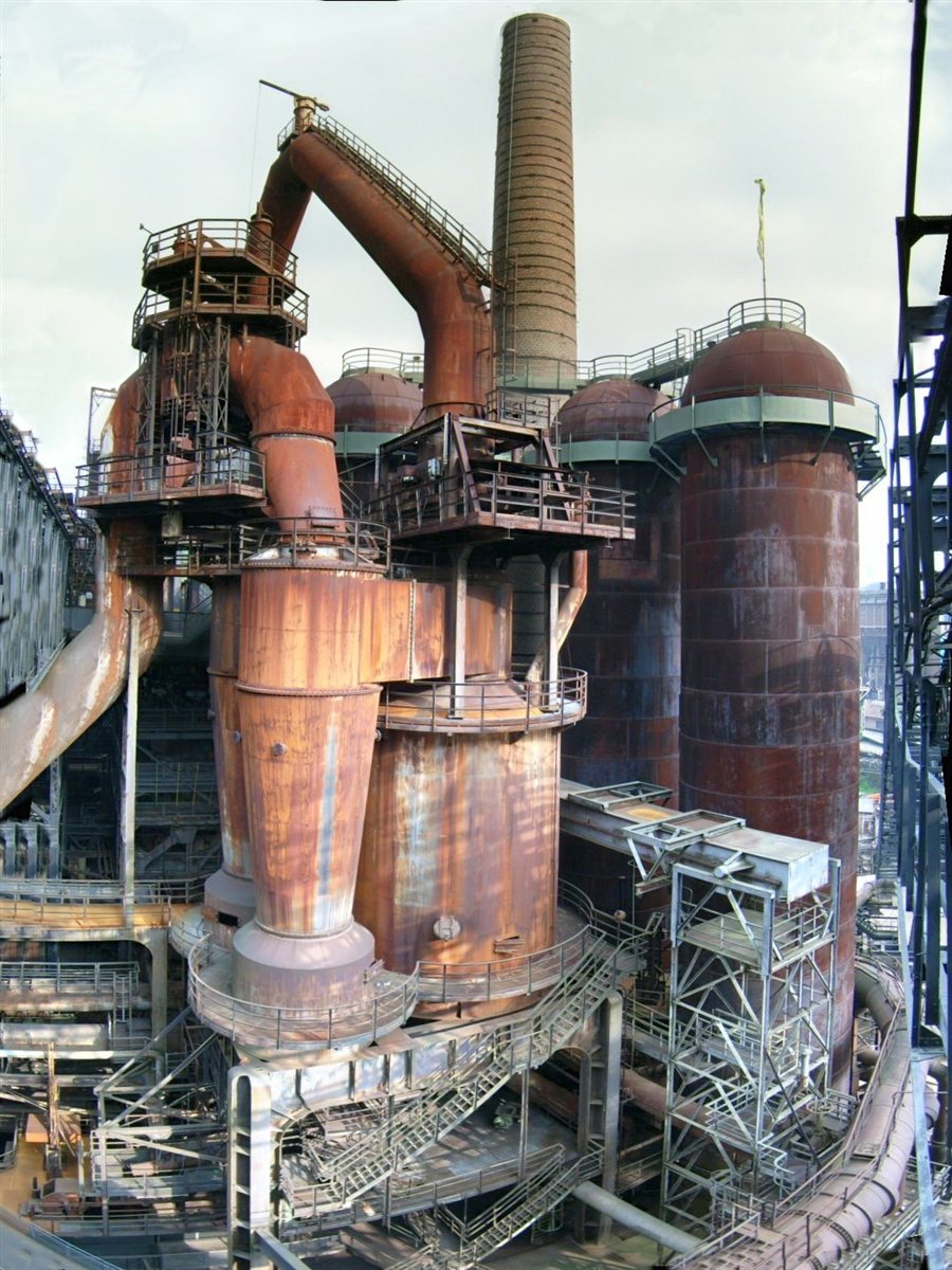 Inside the Völklinger Hütte ironworks: rust-brown blast furnace vessels, domed hot-blast stoves and a tall brick chimney, linked by curving pipework and tiers of steel walkways and staircases