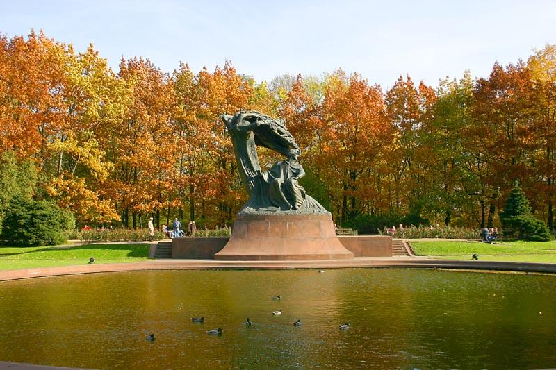 The Chopin monument in Łazienki Park, Warsaw: a bronze figure beneath a windswept willow on a stone plinth, reflected in a pond with ducks, surrounded by trees in autumn colour