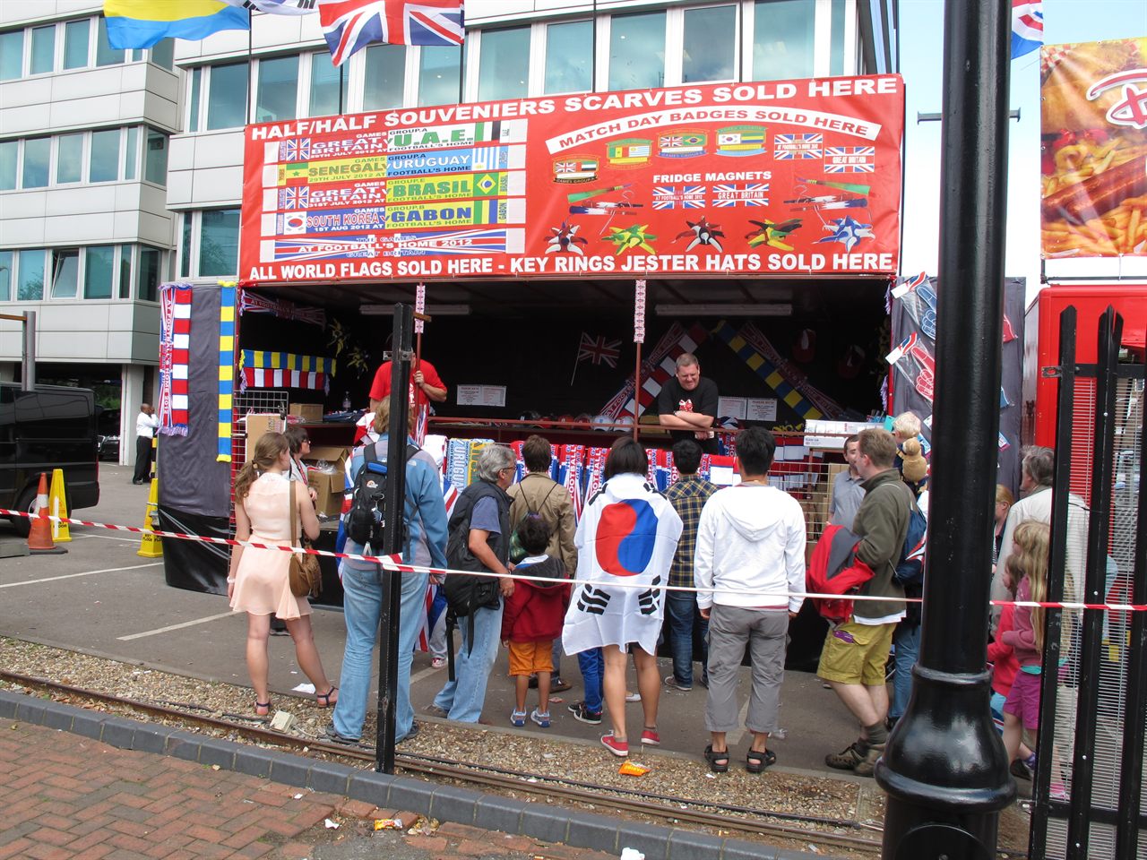 A street stall on the approach to a stadium, hung with dozens of football scarves in club colours, with programmes and flags on the table in front
