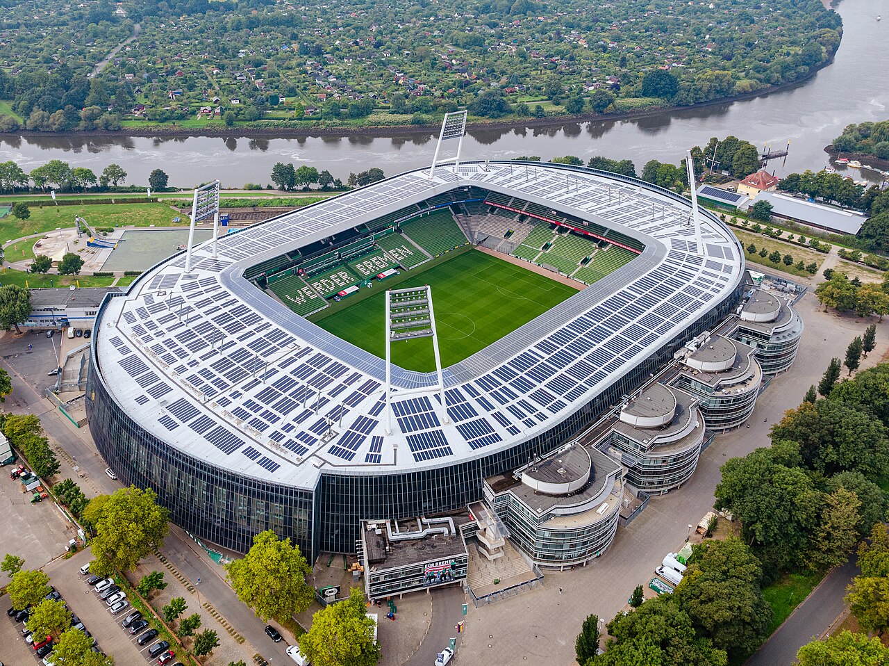 The Weserstadion from the air: an oval stadium whose entire roof and outer facade are covered in dark solar panels, with WERDER BREMEN picked out in the green seats and the river Weser running behind it