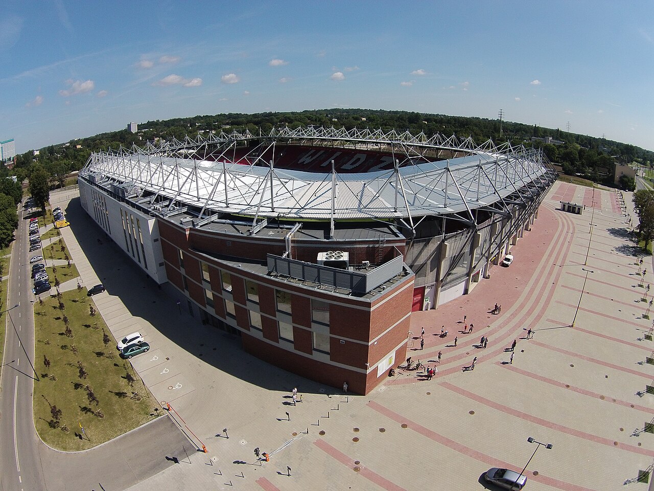 Stadion Widzewa from the air on a summer day: a red-brick and steel stadium with WIDZEW spelled out in the seats of the far stand, a wide paved forecourt in front and woodland behind