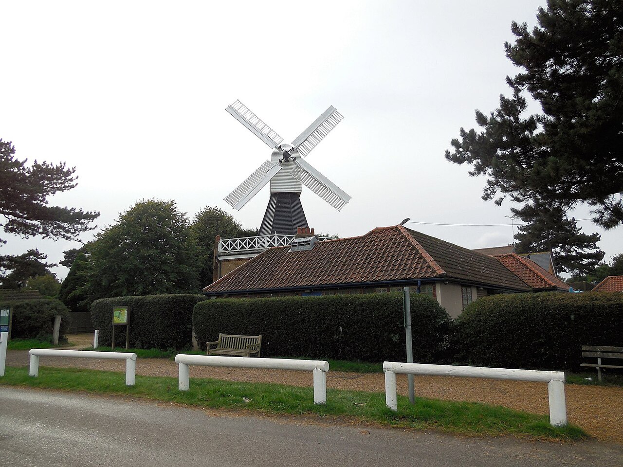 The windmill on Wimbledon Common: a black weatherboarded tower mill with white sails, standing behind a red-tiled building, with clipped hedges, a wooden bench and a white post-and-rail fence in front of it