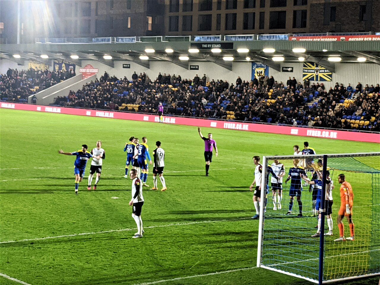A floodlit evening match at Plough Lane: a full stand of blue and yellow seats behind the goal with a large banner reading The Dons Are Back and a Wimbledon flag hanging from it, players in blue and yellow contesting a corner, and apartment blocks rising behind the roof