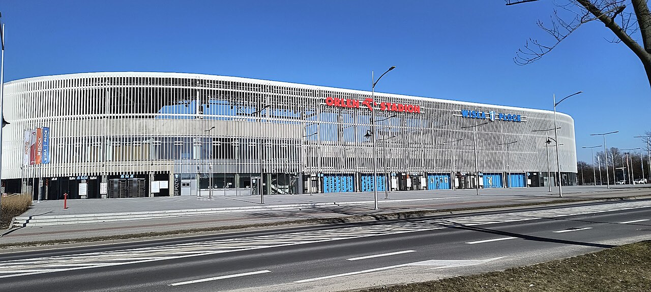 The outside of Wisła Płock's rebuilt stadium: a long curved facade of white vertical louvres with ORLEN STADION in red lettering along it, blue turnstile doors at ground level and an empty road in front