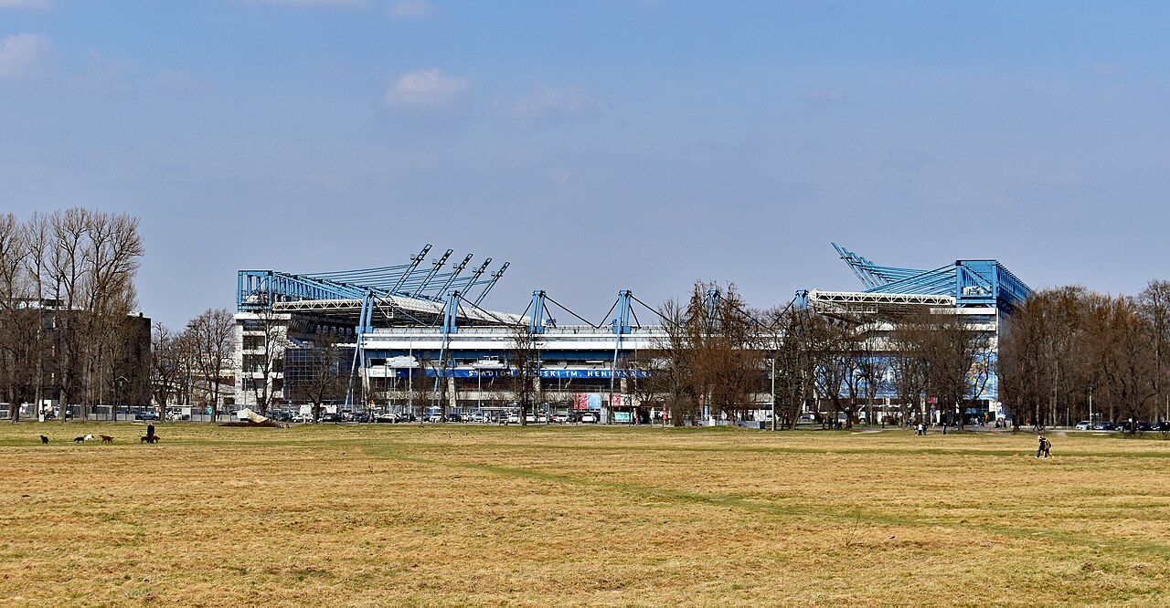 Stadion Miejski im. Henryka Reymana seen across the open grass of the Błonia meadow, its blue floodlight masts and roof trusses rising above bare winter trees