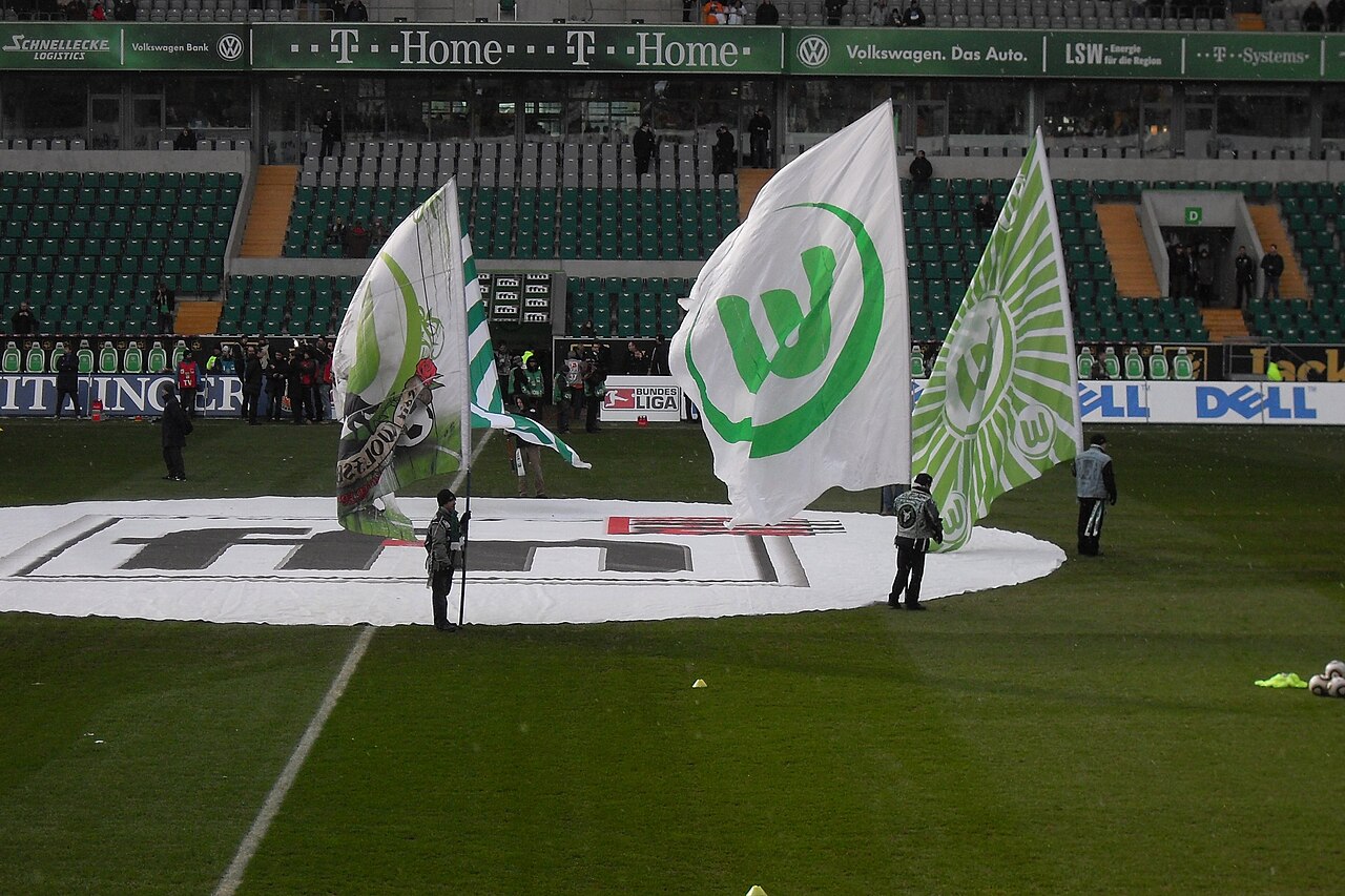 Pitchside at the Volkswagen Arena before kick-off: flag bearers holding large green and white VfL Wolfsburg banners over a circular pitch cover, with Volkswagen advertising along the hoardings and largely empty green and yellow seats behind