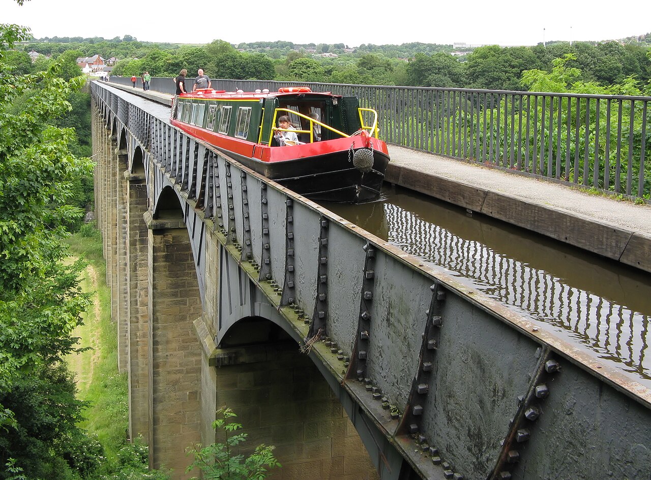 A red and green narrowboat crossing the Pontcysyllte Aqueduct: a cast-iron canal trough carried on tall slender stone piers high above a wooded valley, with a narrow railed towpath alongside and walkers on it