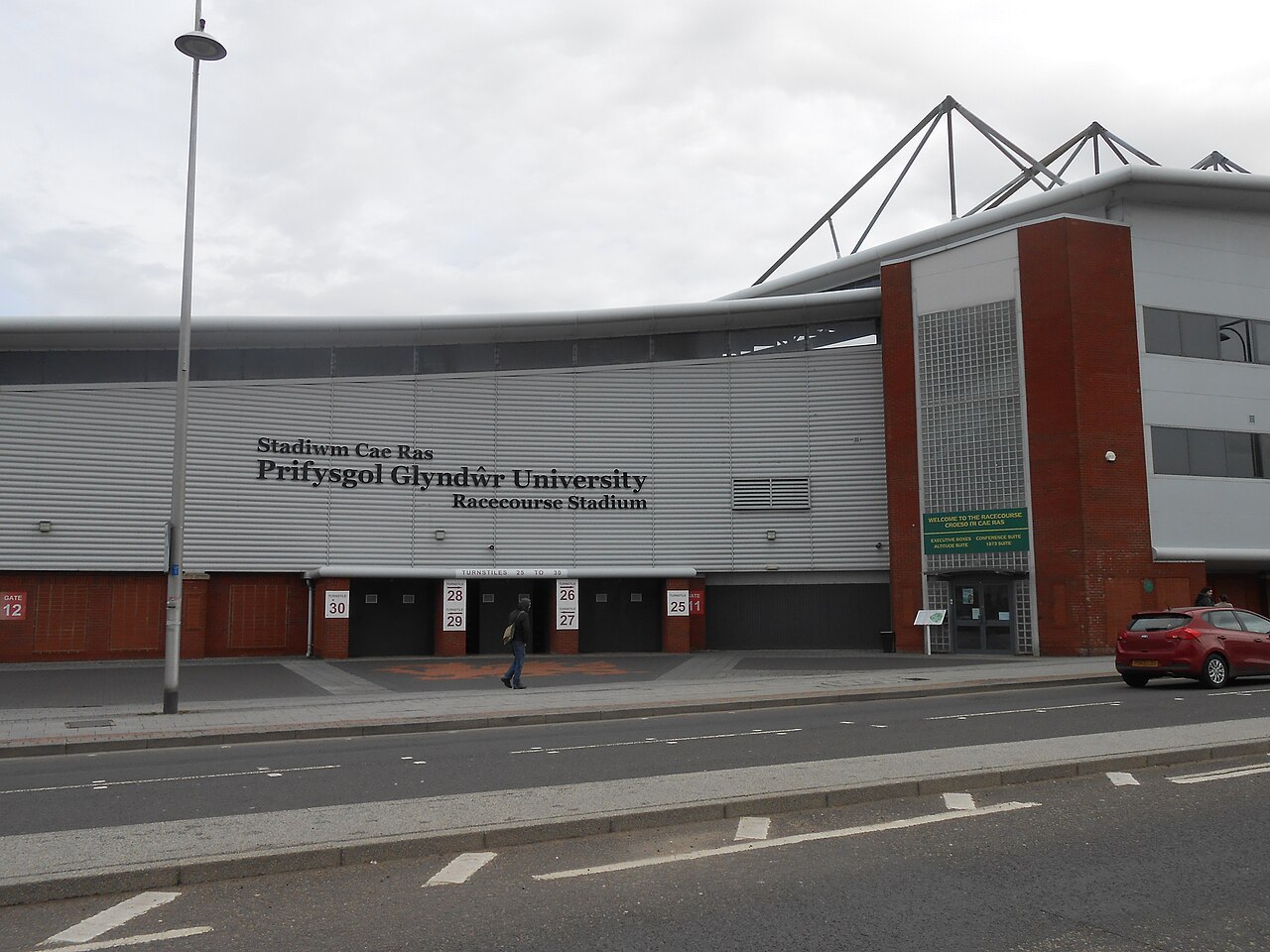 The exterior of the Racecourse Ground in Wrexham from the street: a long silver-clad stand with the bilingual lettering Stadiwm Cae Ras above Racecourse Stadium, a row of turnstiles numbered 25 to 30 along the base, a red brick tower at one end and a supporter walking towards the entrance