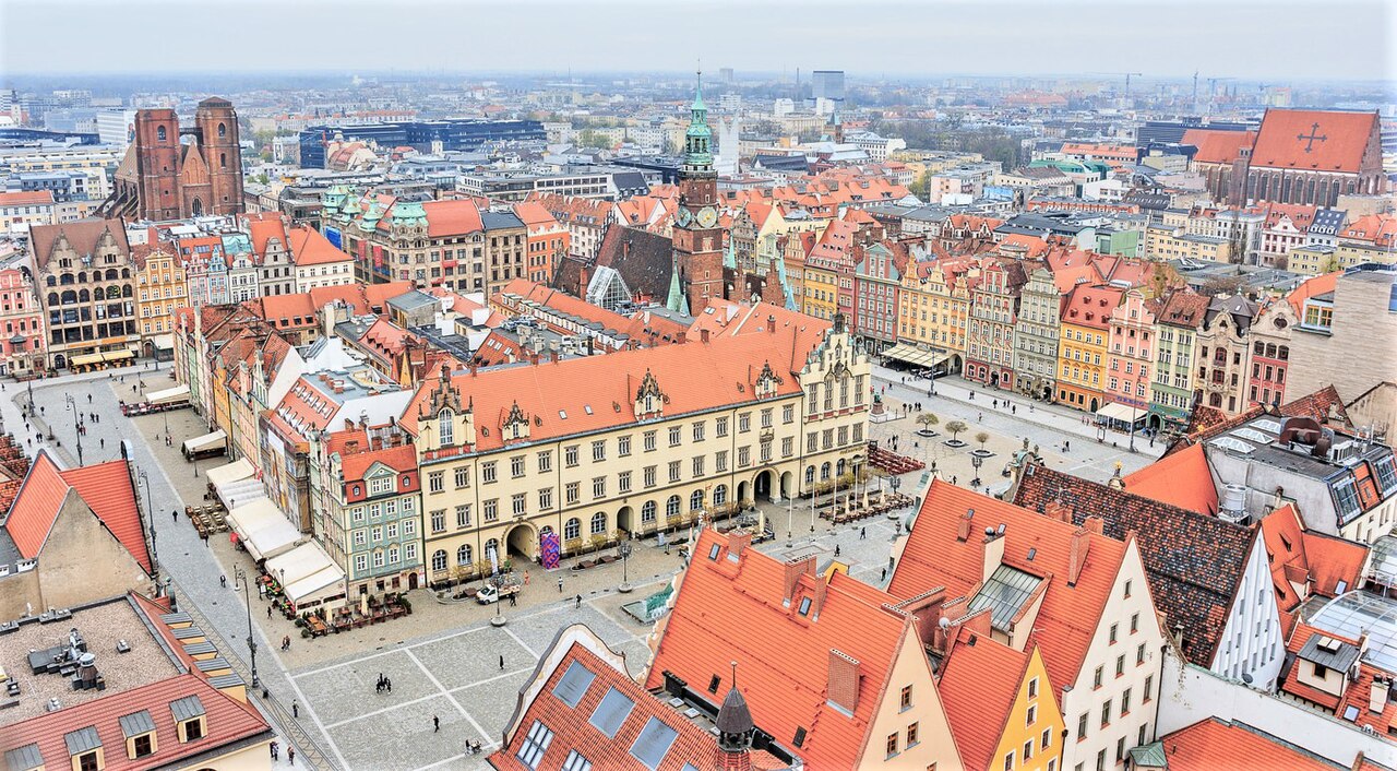 Wrocław's market square from above: the Gothic town hall in the middle of the square, surrounded by rows of tall gabled townhouses in yellow, pink, green and red under orange rooftops