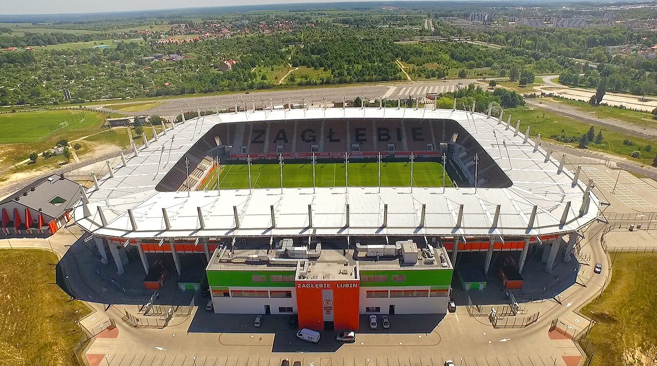 The KGHM Zagłębie Arena from the air: an oval stadium with white roofs over four stands, ZAGŁĘBIE spelled out in the seats of the far side and ZAGŁĘBIE LUBIN on the main stand facade, fields and woodland beyond