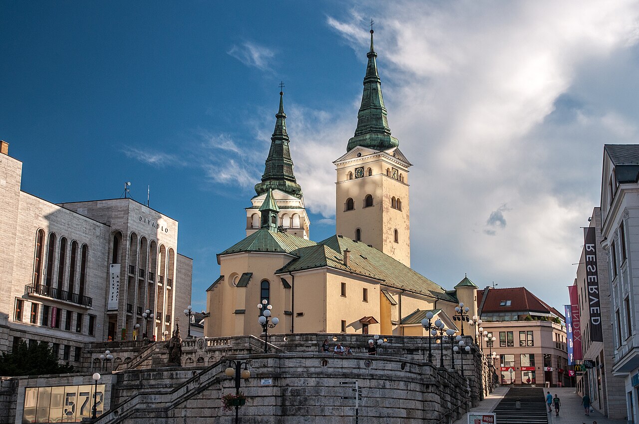 Žilina's Holy Trinity cathedral with two green copper spires above a pale stone terrace of steps, the city theatre on the left and shops along the street on the right