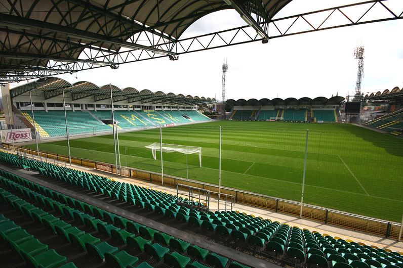 The inside of Štadión pod Dubňom: green seats in four covered stands with MŠK spelled out in the far stand, scalloped white roof canopies and floodlight pylons above an empty pitch